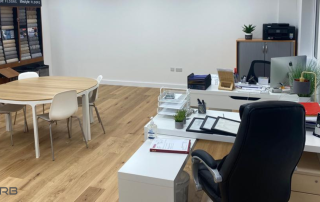 An office after fit out. It shows a laminate floor a pair of desk, a central table with chairs around, and a display unit.